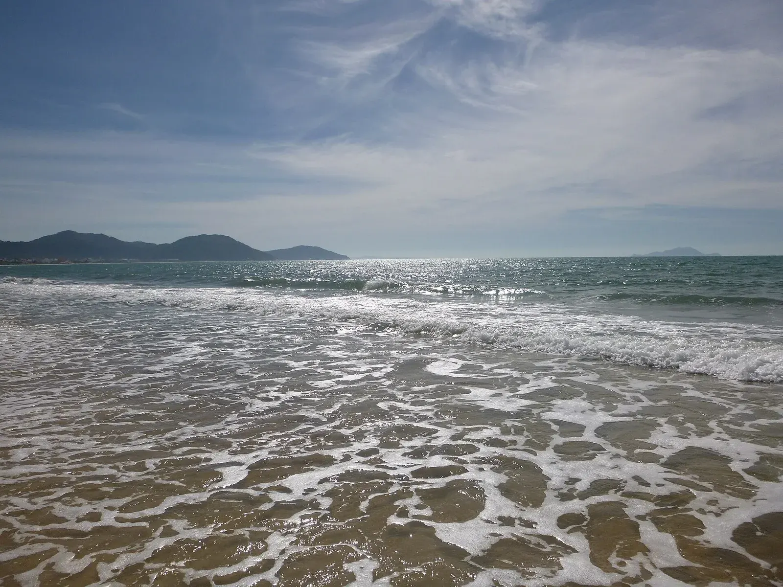 Ondas chegam à areia da Praia do Santinho, entre o mar e os morros.